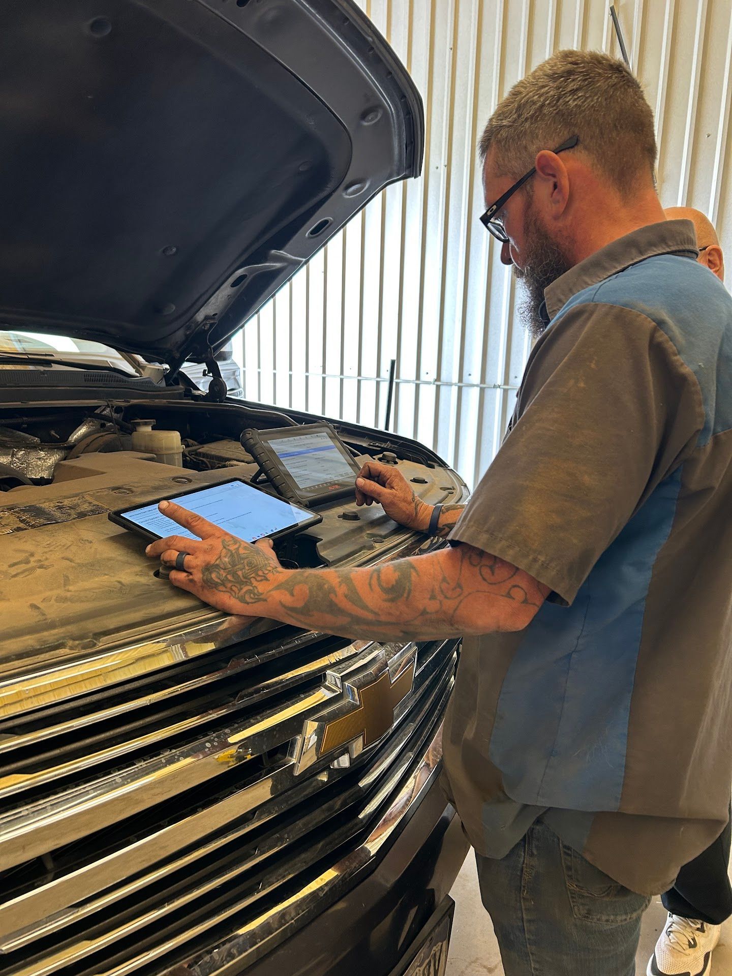 Mechanic using a tablet to diagnose a car with the hood open. Man wears glasses and a work shirt in a garage. | TST Mechanic Services