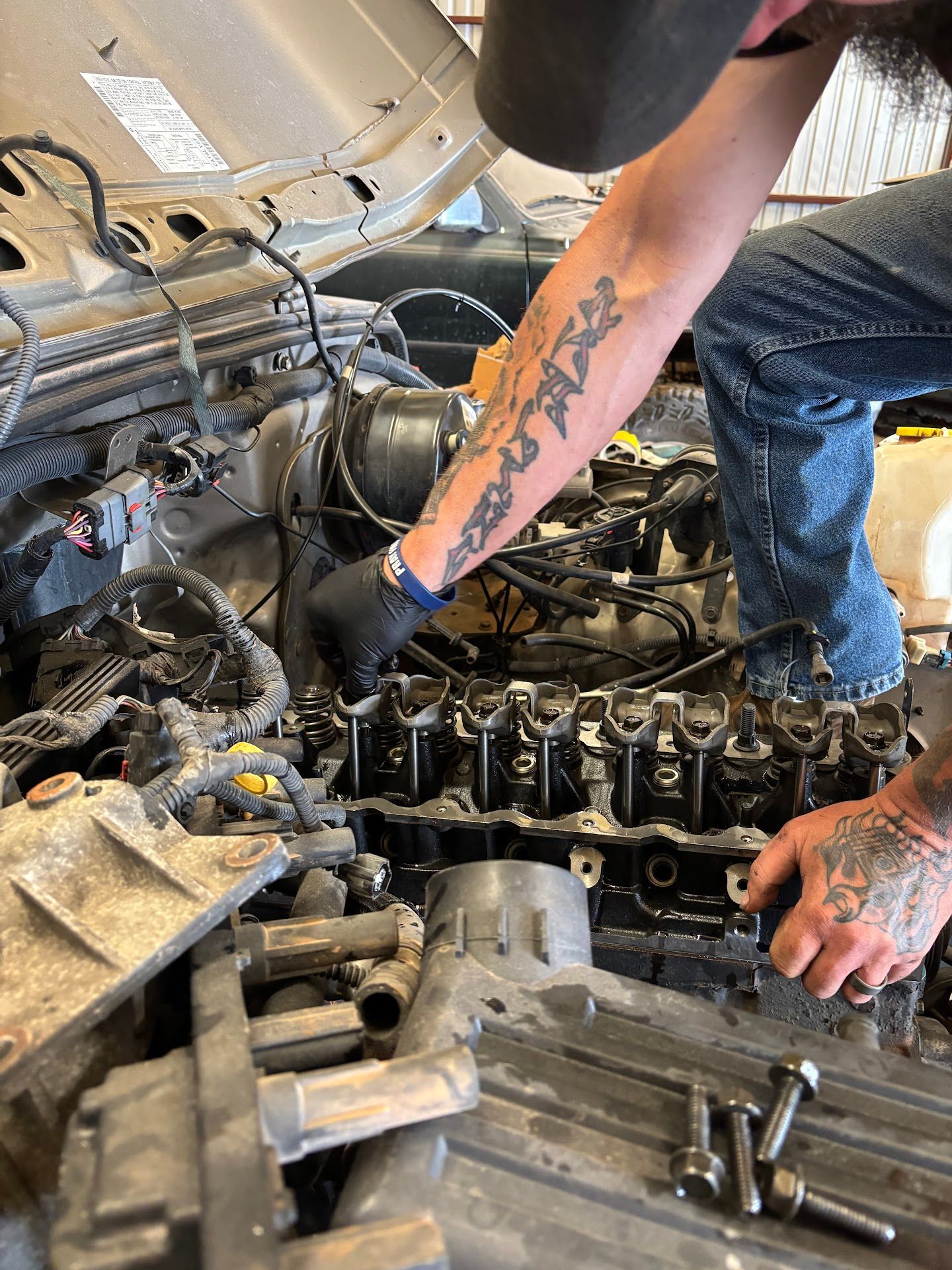 Mechanic working on a car engine; dark gloves, tattoos, visible engine parts, and tools. | TST Mechanic Services