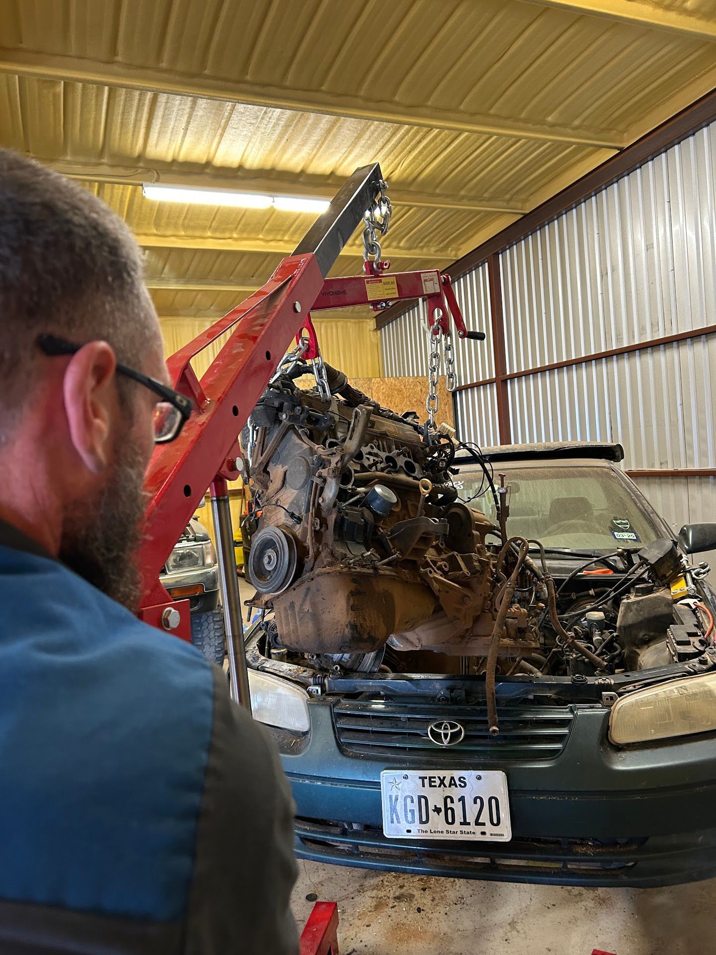 Mechanic watches engine being lifted from green car in a garage. Red crane used. Texas license plate visible. | TST Mechanic Services