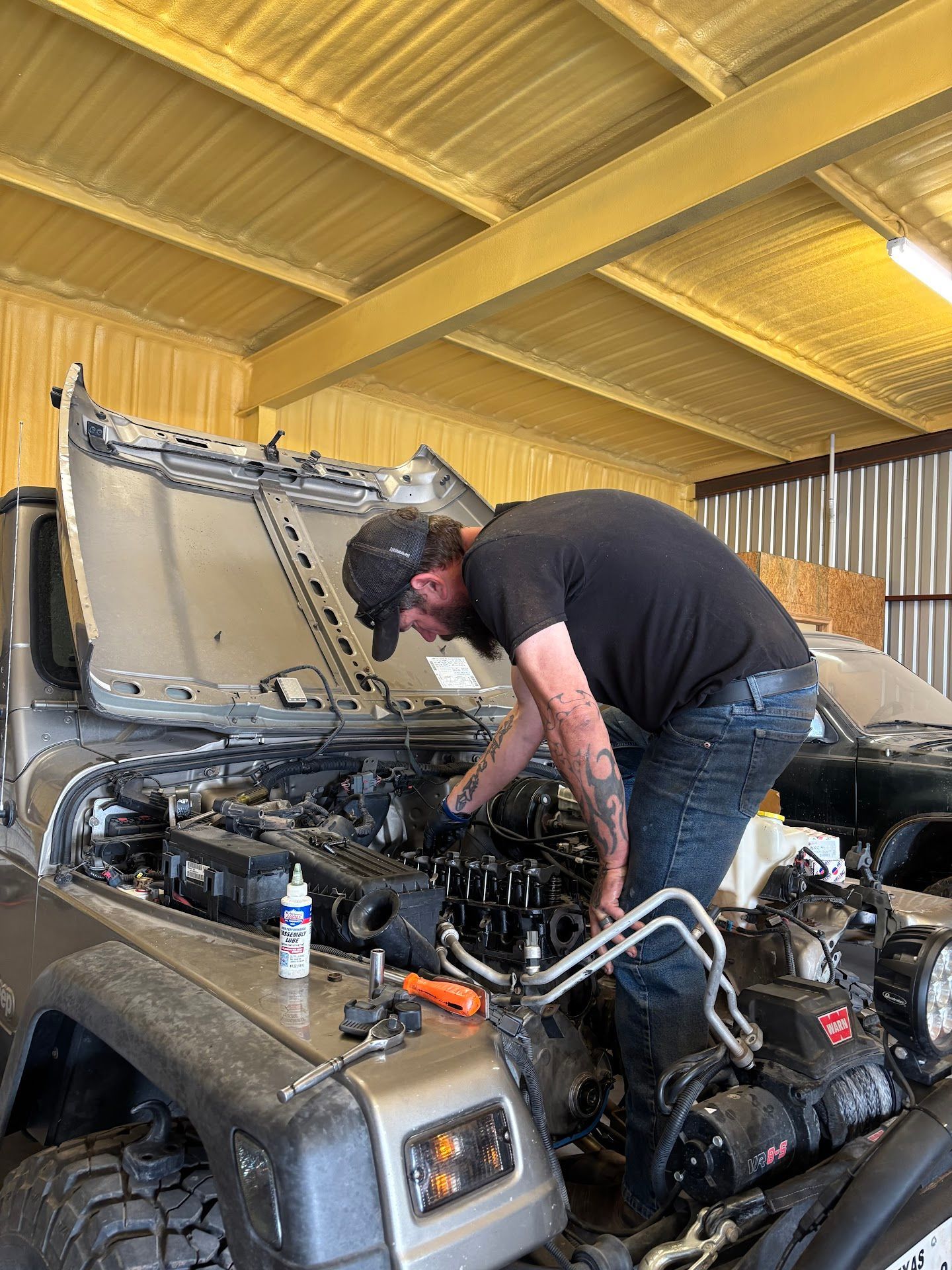 Mechanic working on a Jeep engine, wearing black shirt and jeans, inside a garage. | TST Mechanic Services