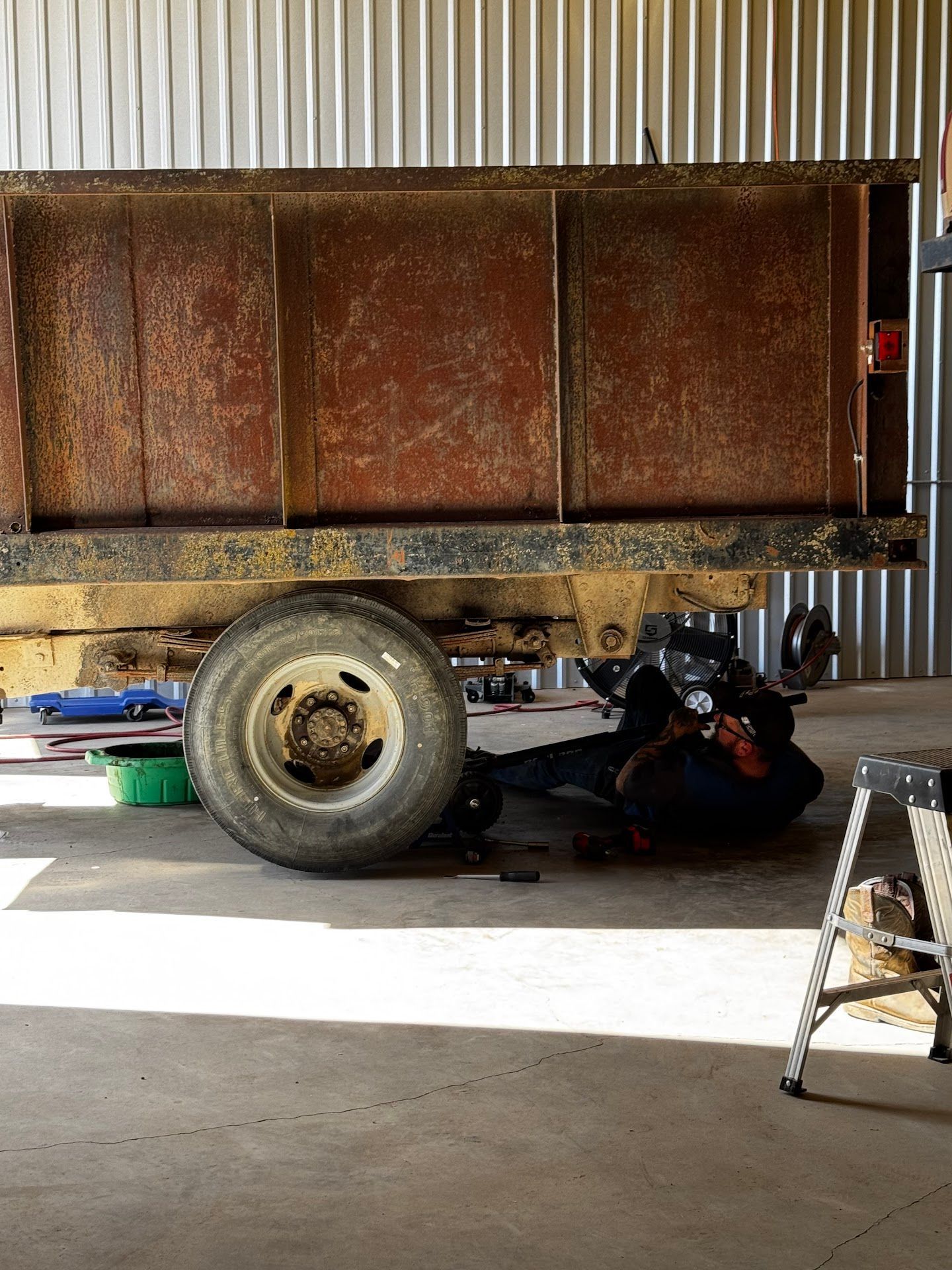 Mechanic working on the underside of a rusty trailer in a garage, with a green bucket and small ladder present. | TST Mechanic Services