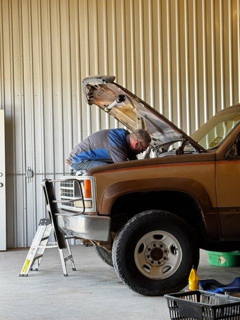 Mechanic working on a brown truck in a garage, hood open. Standing on the bumper, uses a stepladder. | TST Mechanic Services