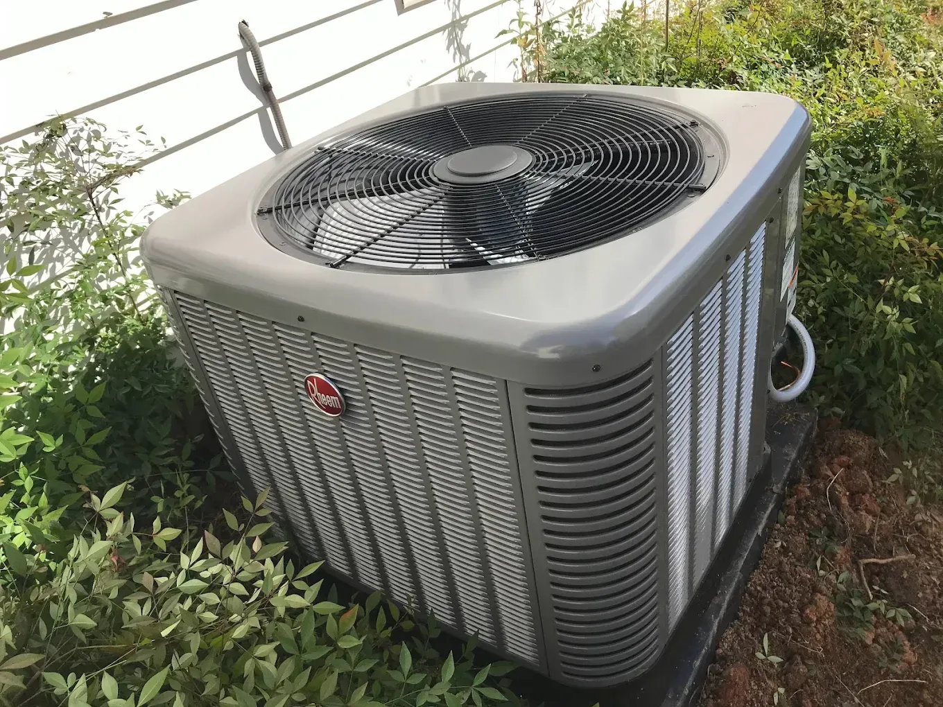 A gray Rheem air conditioning unit outdoors near a white wall and green bushes.