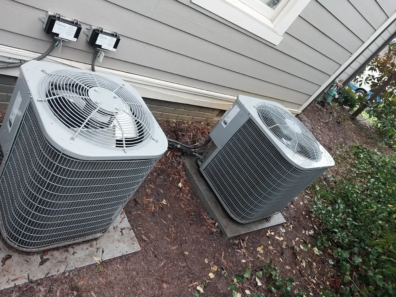 Two air conditioning units outside a building on gravel, near foliage. Gray and white colors.