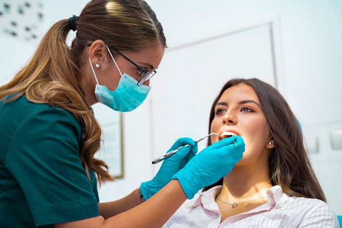 Dentist examining a patient's teeth. The dentist, wearing a mask and gloves, holds tools while the patient's mouth is open.