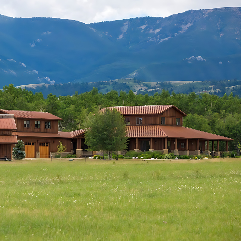 A large house sits in the middle of a grassy field with mountains in the background