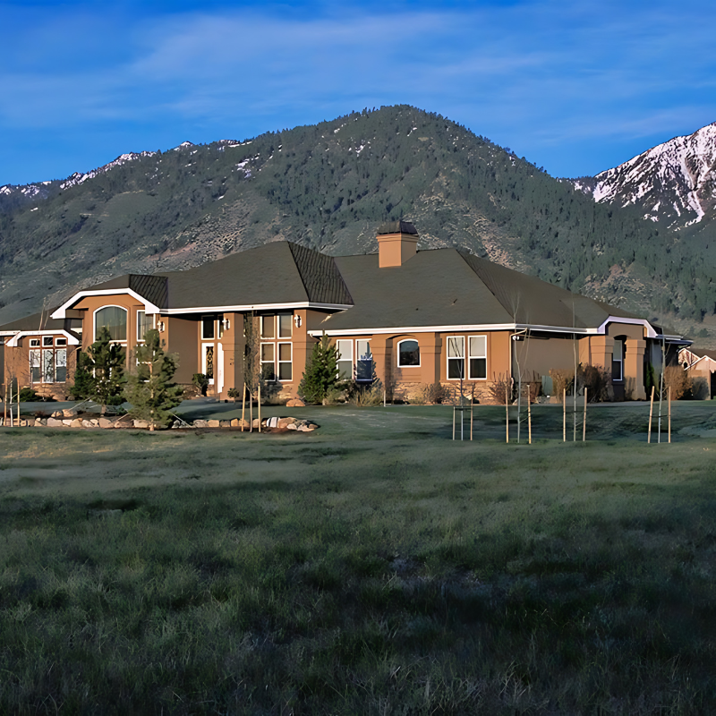 A large brick house sits in the middle of a grassy field with mountains in the background