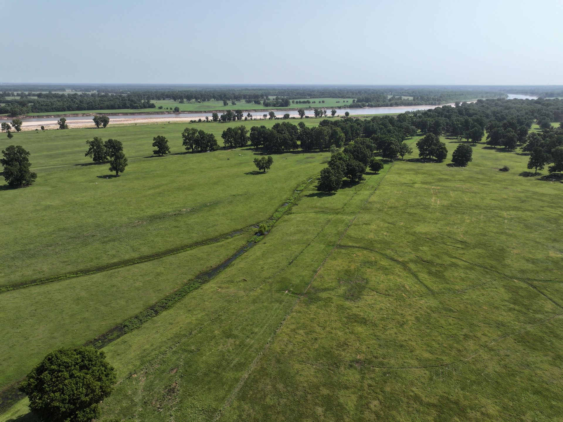 An aerial view of a grassy field with trees and a river in the background