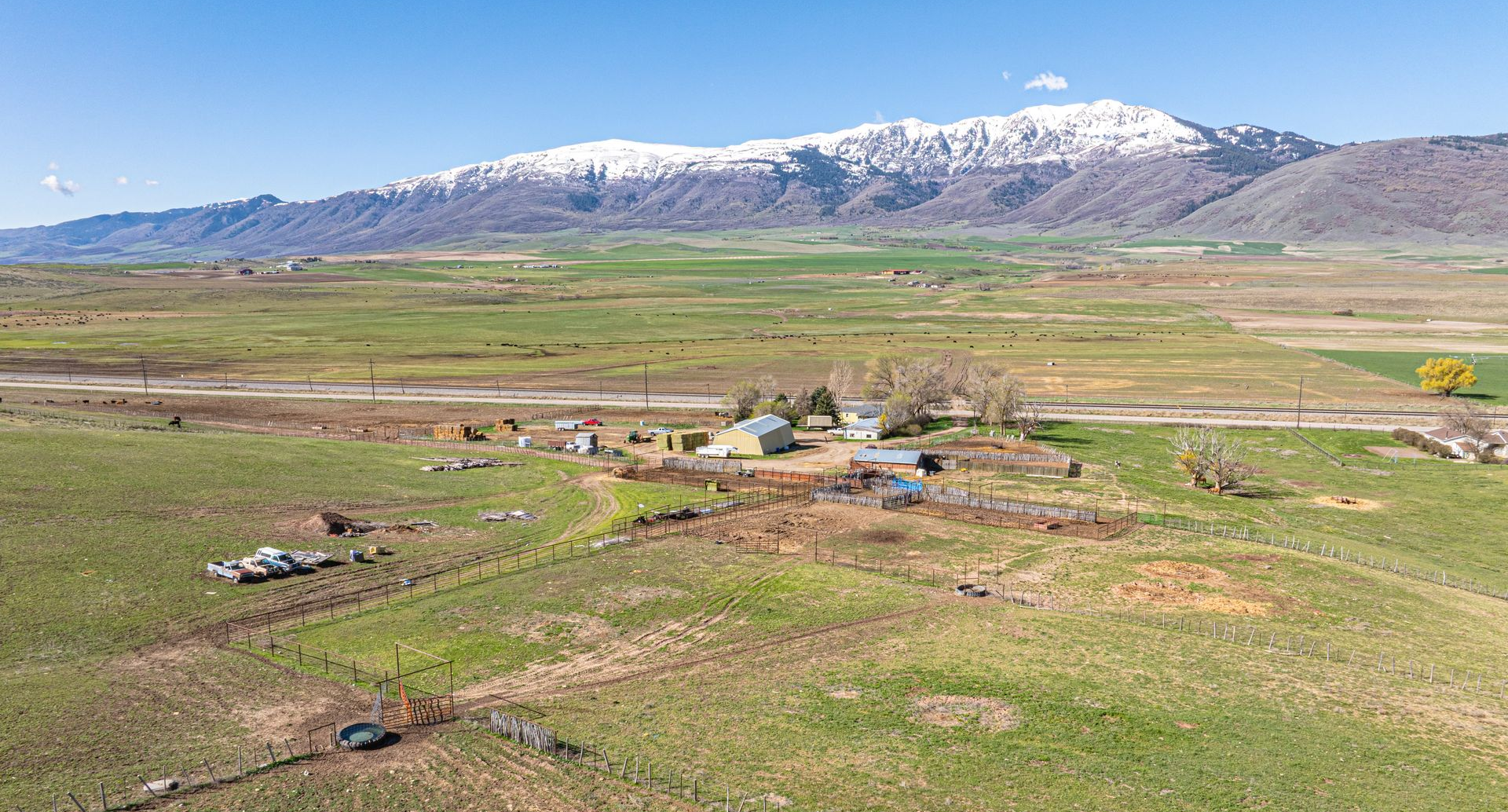 A farm with several small buildings sits in a vast, grassy landscape beneath snow-capped mountains on a sunny day.