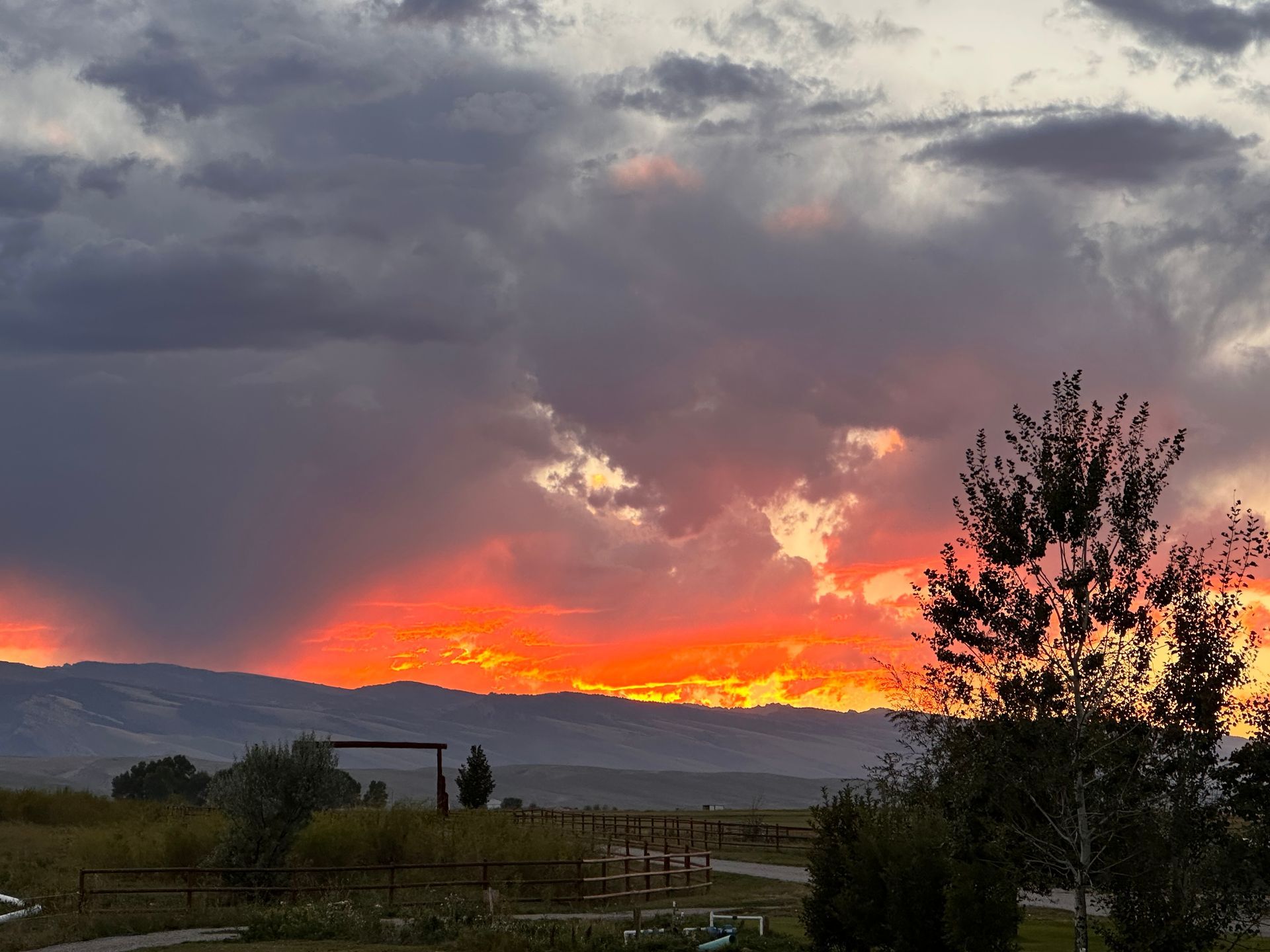Vibrant sunset with fiery orange light glowing behind dark, dramatic clouds over a distant mountain range and rural field.