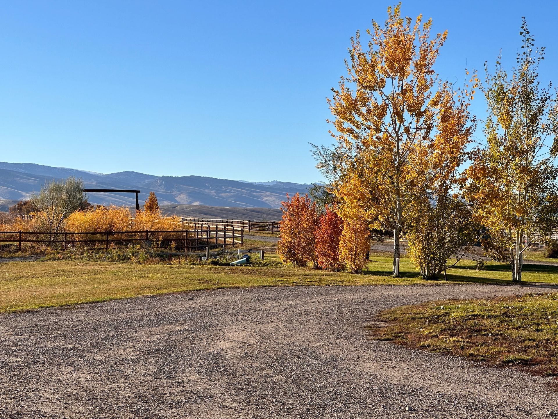 A gravel driveway leads toward a wooden fence and trees with yellow autumn leaves, set against distant mountain ridges.