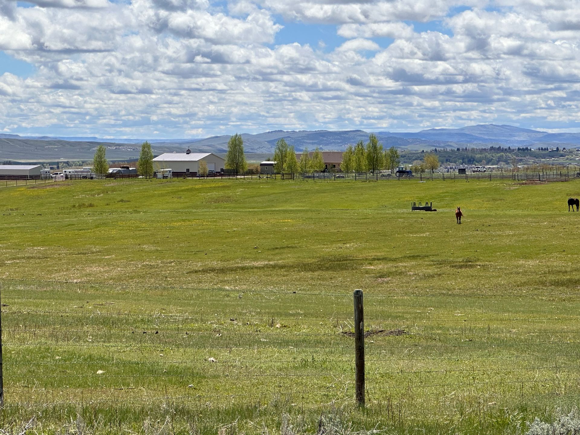 A grassy field under a cloudy blue sky with a fence in the foreground and farm buildings and mountains in the distance.