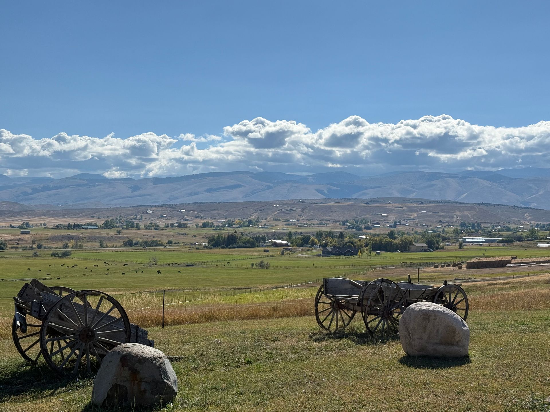 Green farmland and structures with mountains in the background under a blue sky.