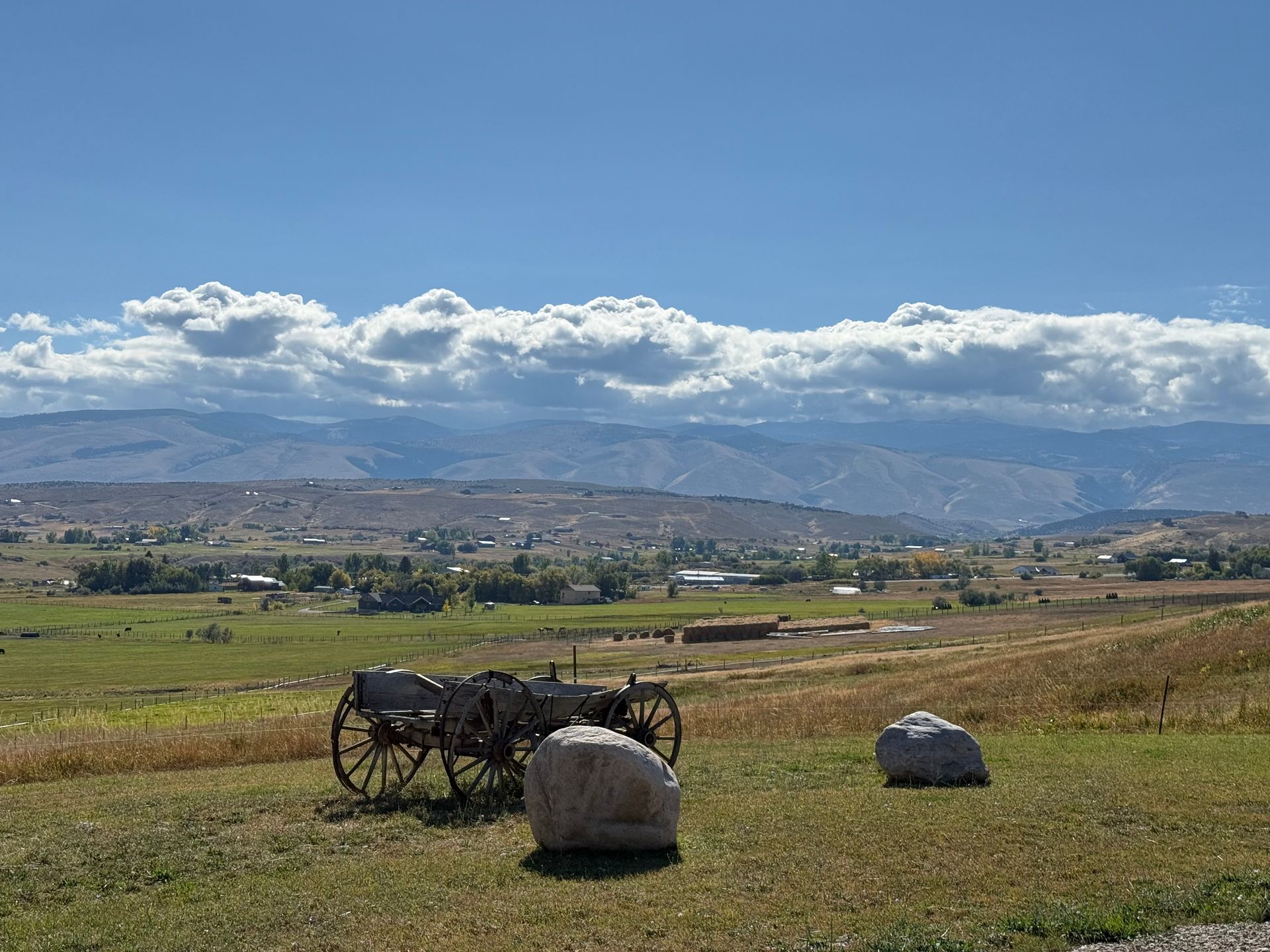 An old wooden wagon sits on a grassy hill overlooking a valley with rolling mountains under a bright, cloudy blue sky.
