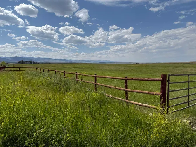 A brown wooden fence runs through a grassy, open field under a blue sky with scattered clouds and distant mountains.