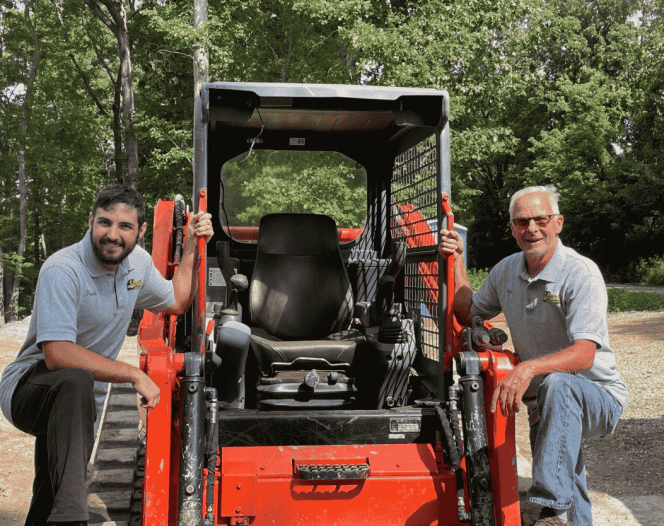 Two men by an orange skid steer loader in a wooded area. The man on the left is kneeling and smiling.