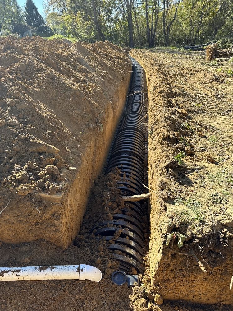 Trench dug with black corrugated drainage pipe installed, ready for backfill. Outdoor, daylight.