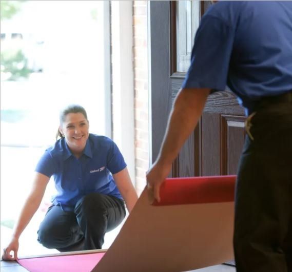 Two movers placing a red carpet inside a doorway. Woman kneels, smiling; man holds rolled carpet.