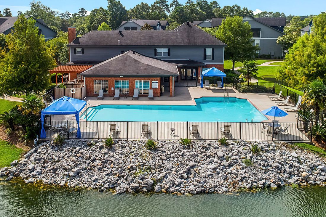Pool area with building and lake in background. Blue cabanas, lounge chairs, and stone seawall.