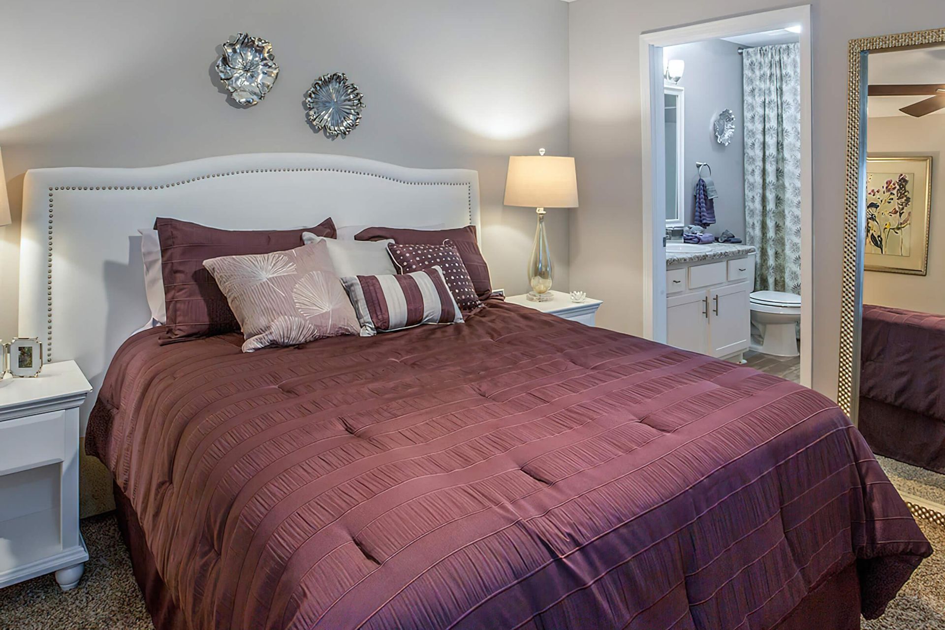 Bedroom with burgundy bedding, white headboard, and adjacent bathroom visible in a large mirror.
