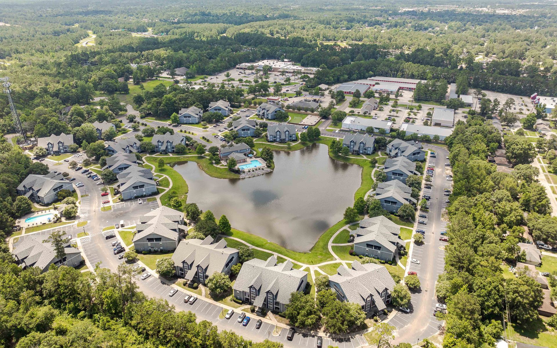 Aerial view of apartment complex with a lake, surrounded by trees and a shopping center.