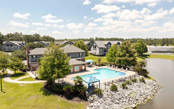 Aerial view: Pool and clubhouse next to a pond at an apartment complex. Blue sky, green trees, and buildings.