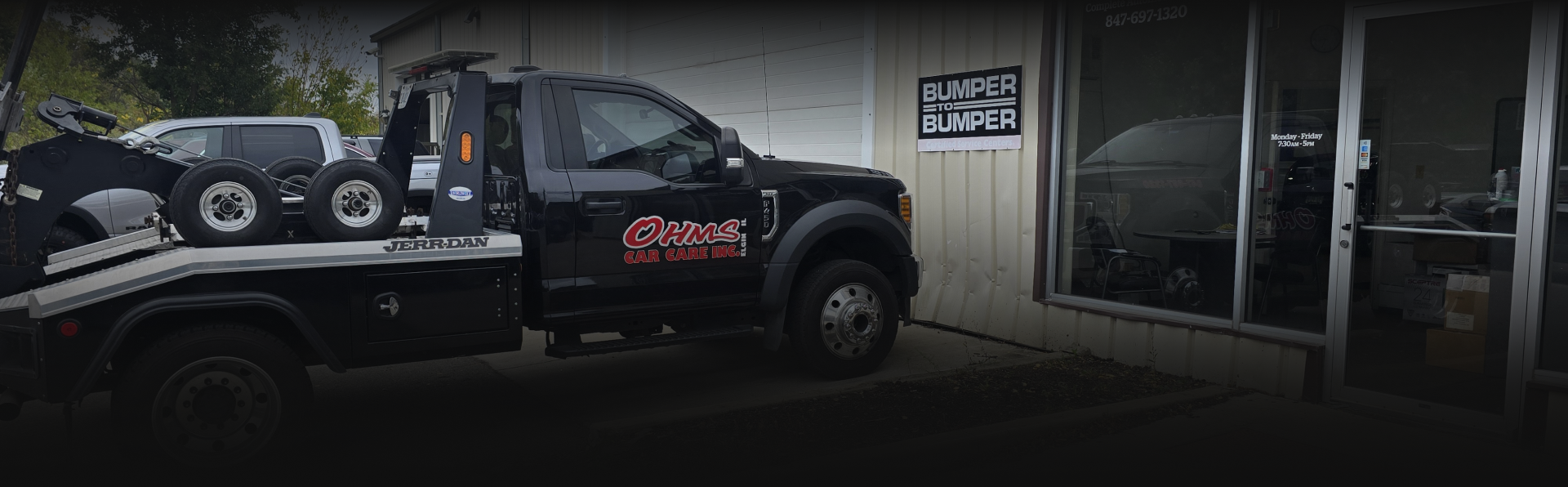 Black tow truck in front of a shop, tires on the bed. Shop sign visible - OHMS Car Care & TOWING
