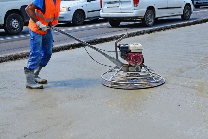 Worker in orange vest floats fresh concrete on a road, cars in background.