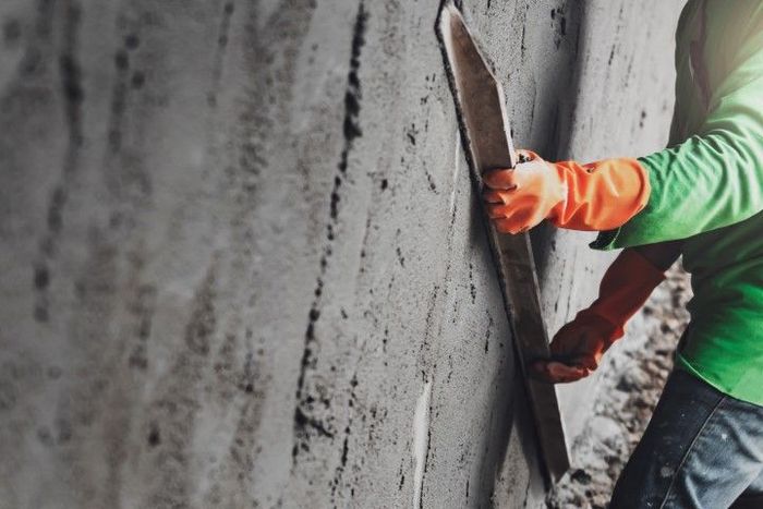 Person in gloves smoothing wet cement on a wall with a long trowel.