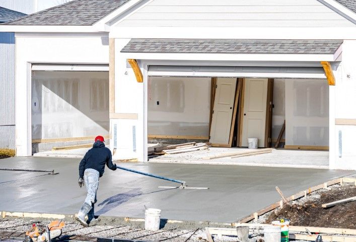 Man smoothing wet concrete in front of a garage under construction; blue jacket, red hat, sunny day.