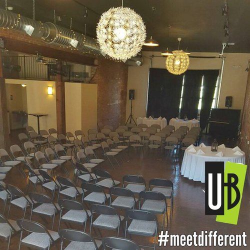Rows of chairs in a circle face a stage with tables in an open room. Two ornate lights hang.