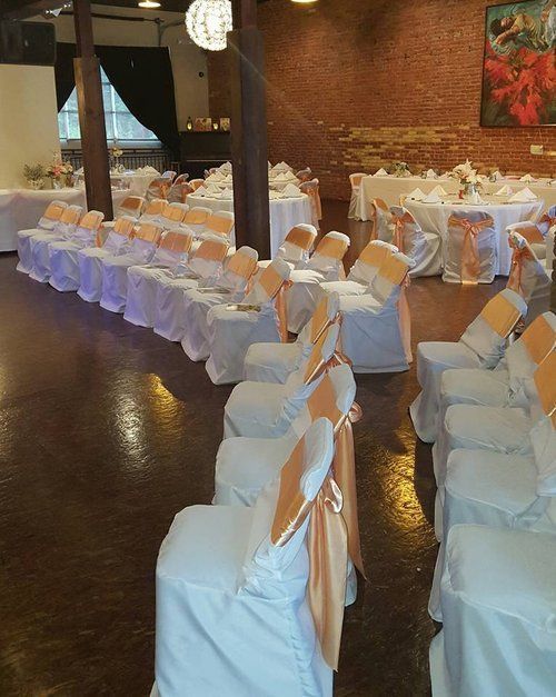 Wedding reception setup with white tablecloths, peach chair sashes, and brick wall backdrop.