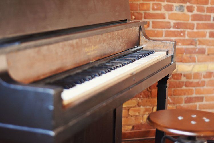 Upright piano in a room with a brick wall and a wooden stool.