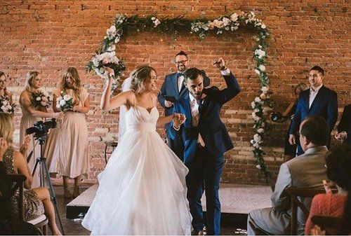 Newly married couple dances, arms raised, at the altar decorated with flowers, in front of a brick wall.