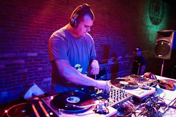 DJ at a club, mixing music on turntables, wearing headphones. Purple and blue lighting, brick wall background.