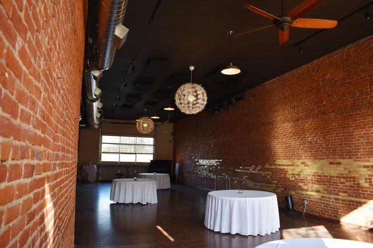 Empty event space with exposed brick walls, round tables draped in white, and decorative hanging lights.