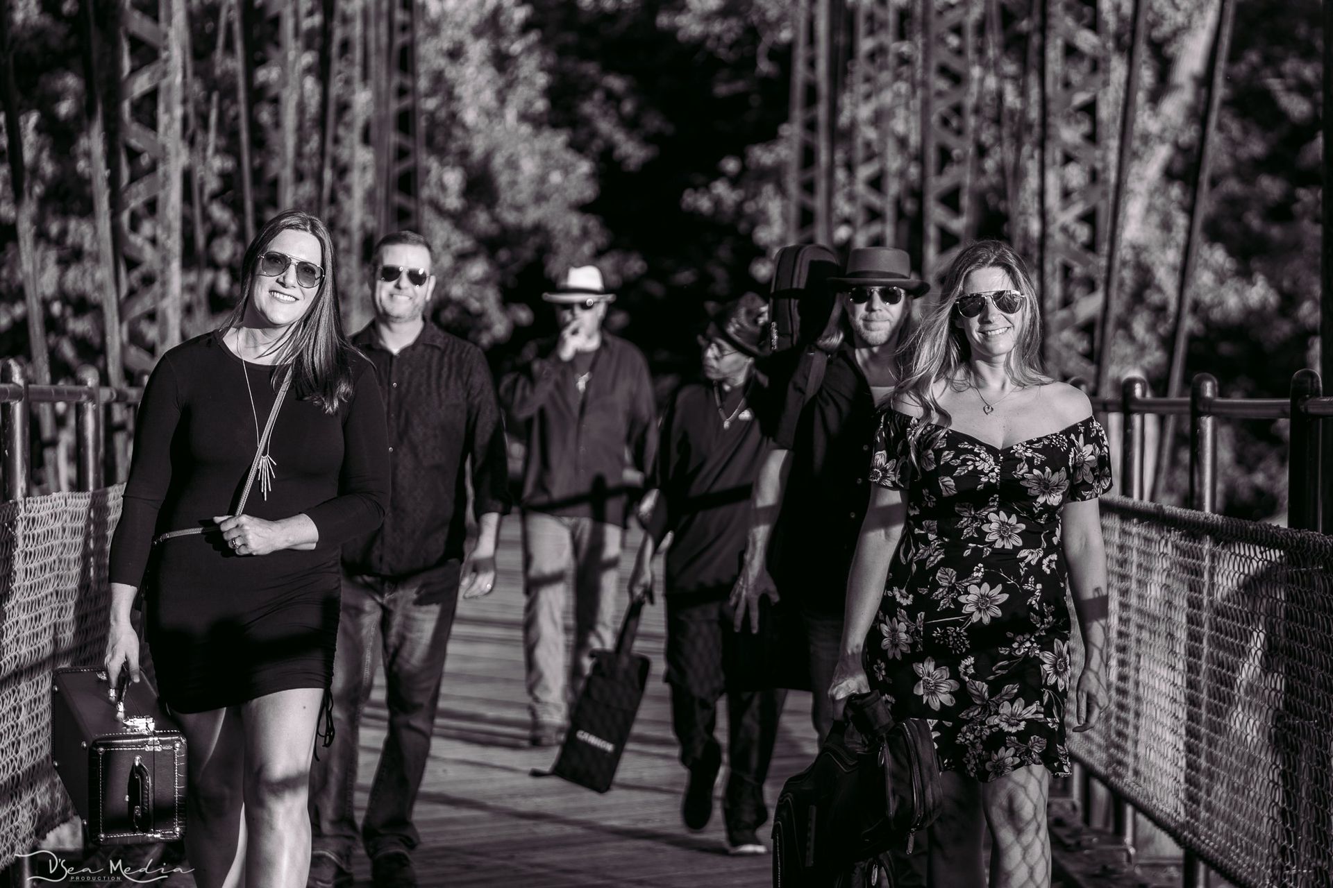 Group walking on a metal bridge. They are in black and white. One carries a guitar.