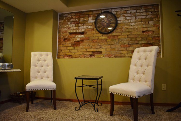 Two white button-tufted chairs flanking a small table; brick wall backdrop with clock; olive green walls and carpet.