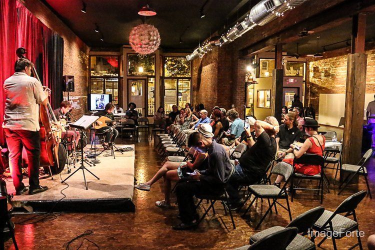 Jazz band performing onstage in a venue with an audience seated at tables, brick walls, and dim lighting.