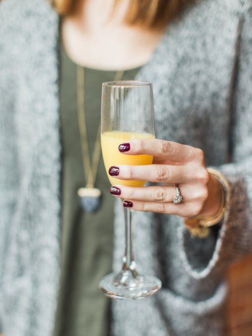 Woman holding a champagne flute filled with orange juice, wearing a grey sweater, with maroon nail polish.