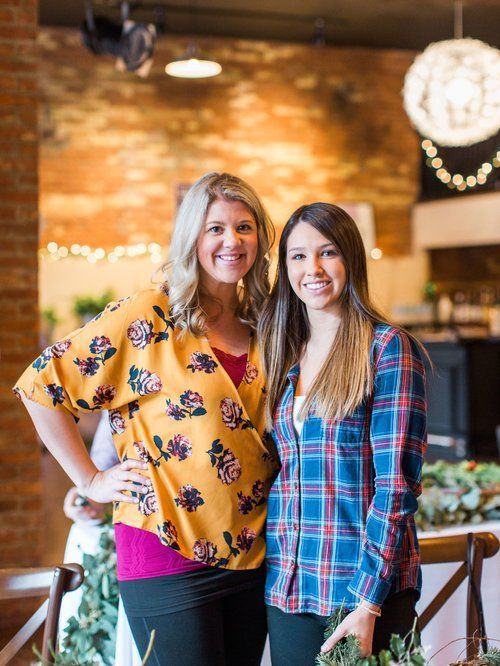 Two women smiling in a room; one in a floral top, the other in a plaid shirt.