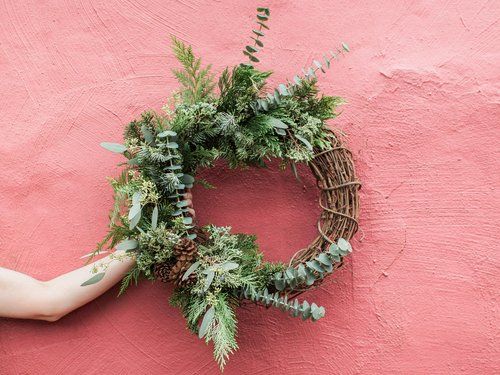 Wreath of greenery and pinecones hangs on a pink wall. A hand holds the wreath.