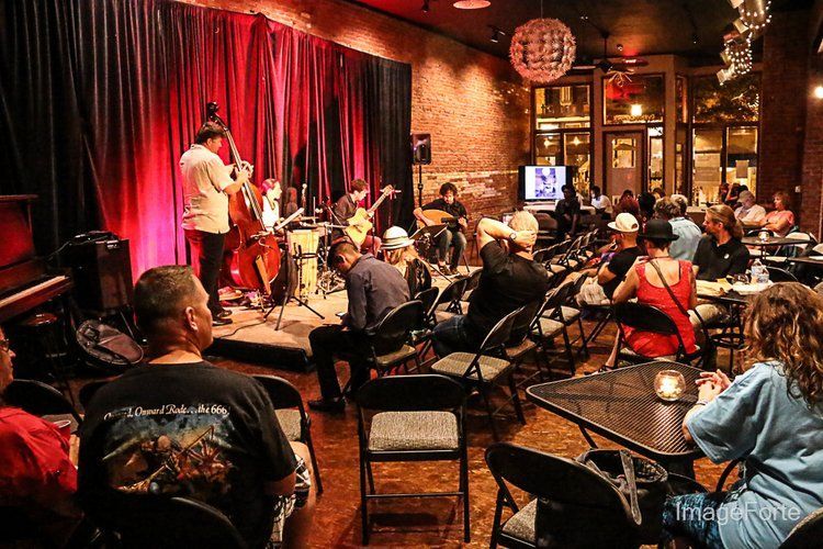 Jazz band performing on stage in a dimly lit club. Audience seated at tables, watching the performance. Red curtains in the background.