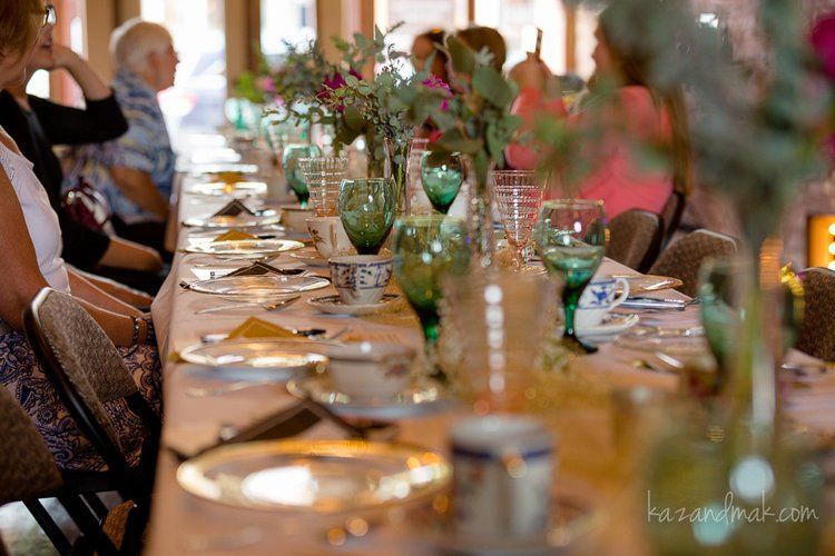 Long table set for a tea party with flowers, vintage glassware, and people seated.