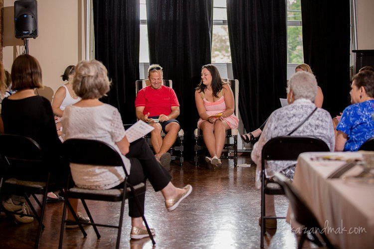 People in chairs, gathered in a room, conversing. Two individuals in the center are talking.