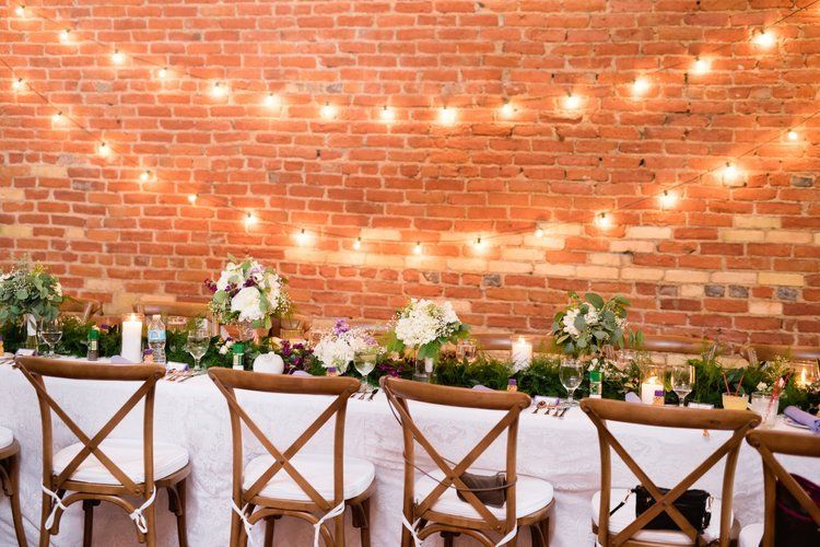 Reception table set against a brick wall, strung with lights. Floral centerpieces, cross-back chairs.