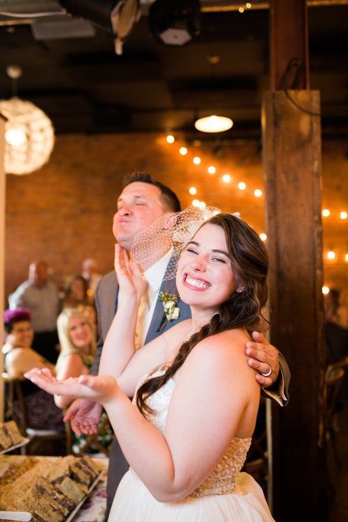 Bride playfully wiping cake off groom’s face during a wedding reception, brick wall and string lights in background.