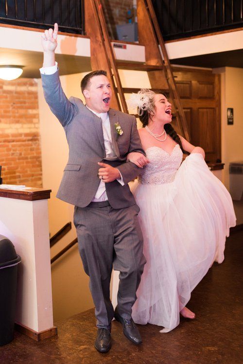 Newlyweds enter a reception. The groom raises arm, bride laughs, white dress flowing. Brick walls, wooden staircase.