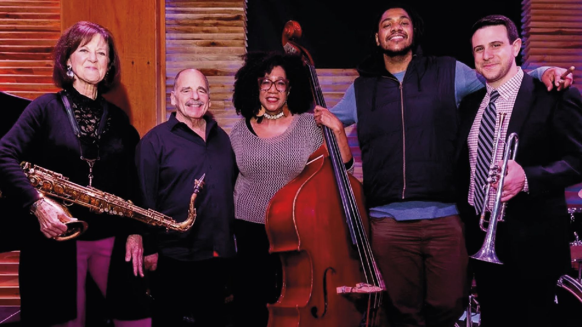 Jazz band poses: saxophone, trumpet, bass, and two musicians smile in a studio setting with a wooden background.