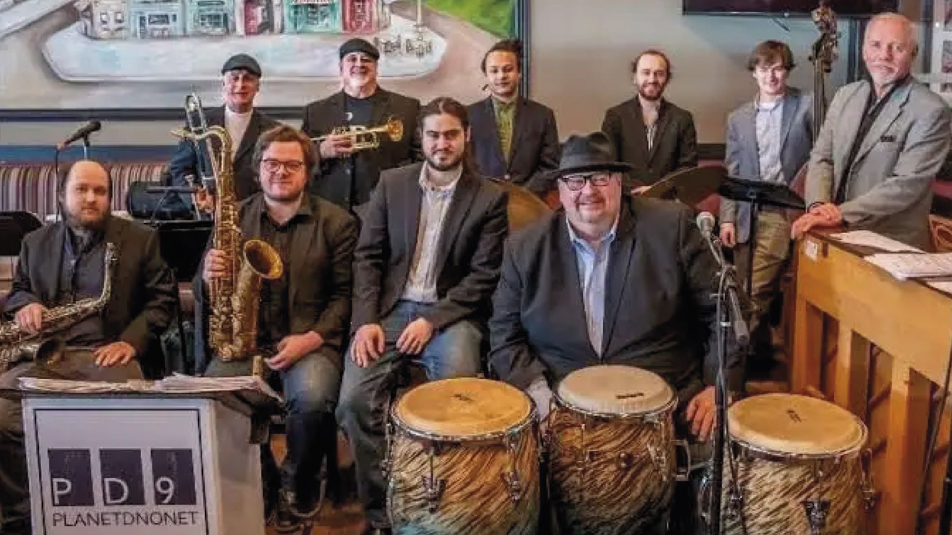 Band Planetonomik, nine men pose together, some with instruments, in front of a window.